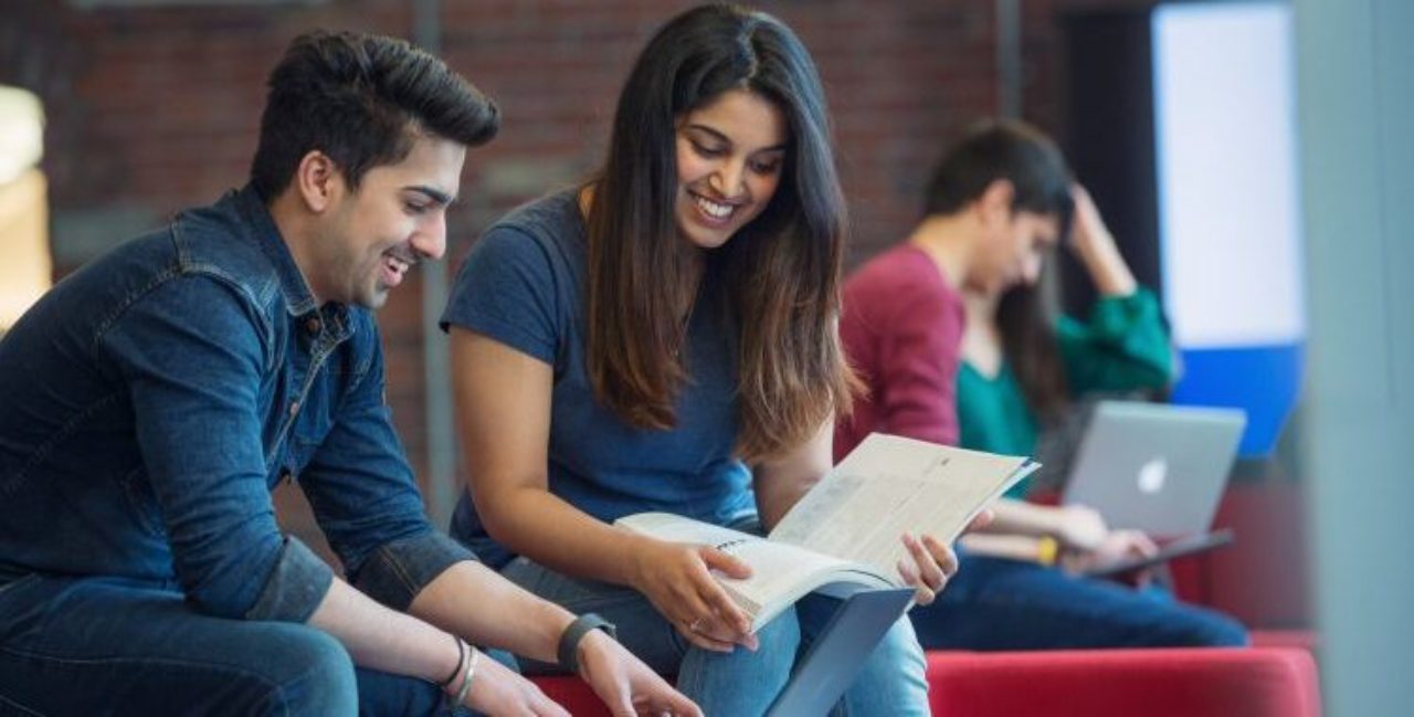 two FDU students smiling while sharing a textbook and working on a laptop