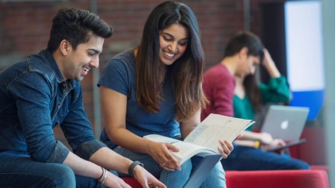 two FDU students smiling while sharing a textbook and working on a laptop