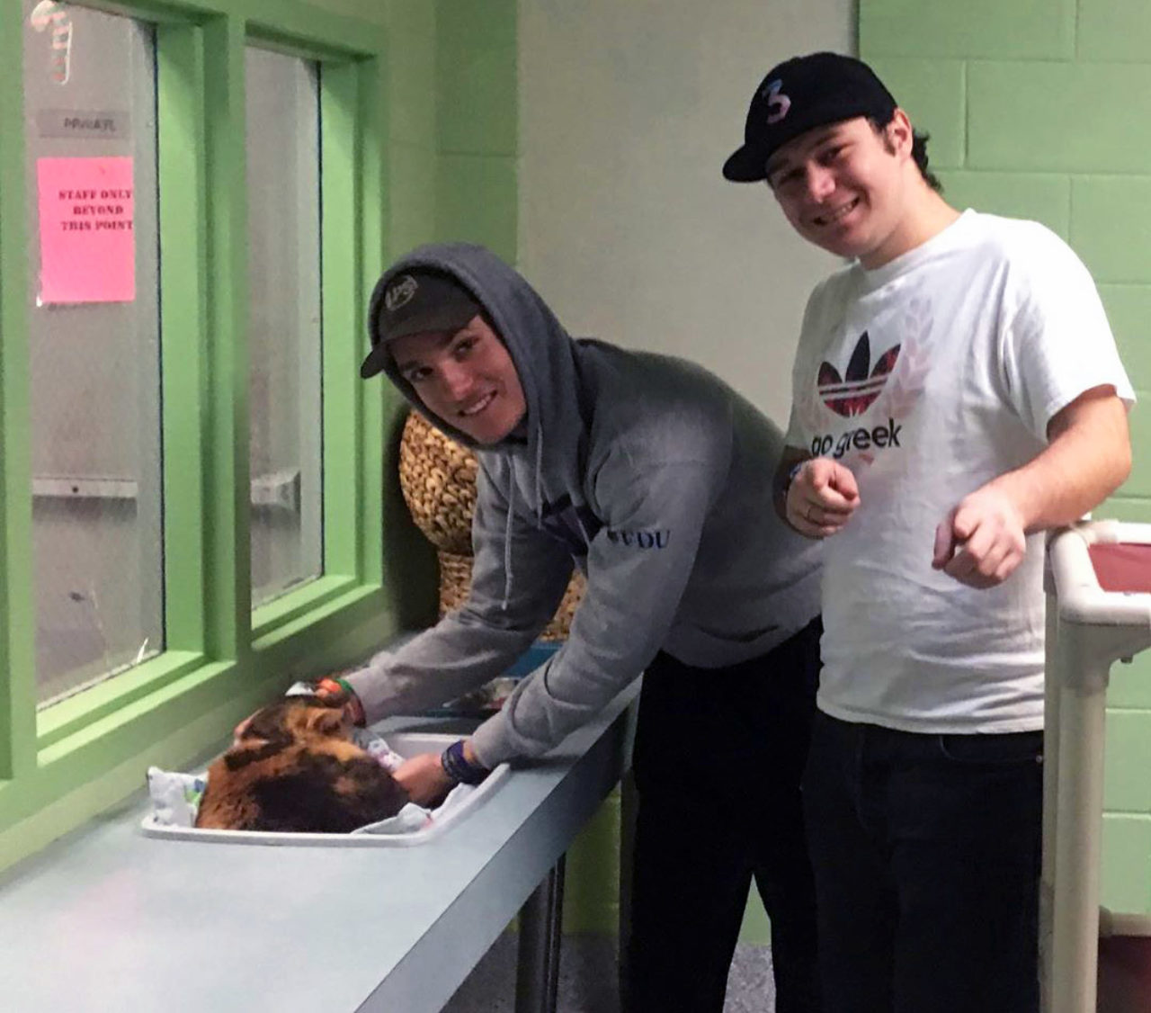 One student pets a cat while the other smiles and points at the camera
