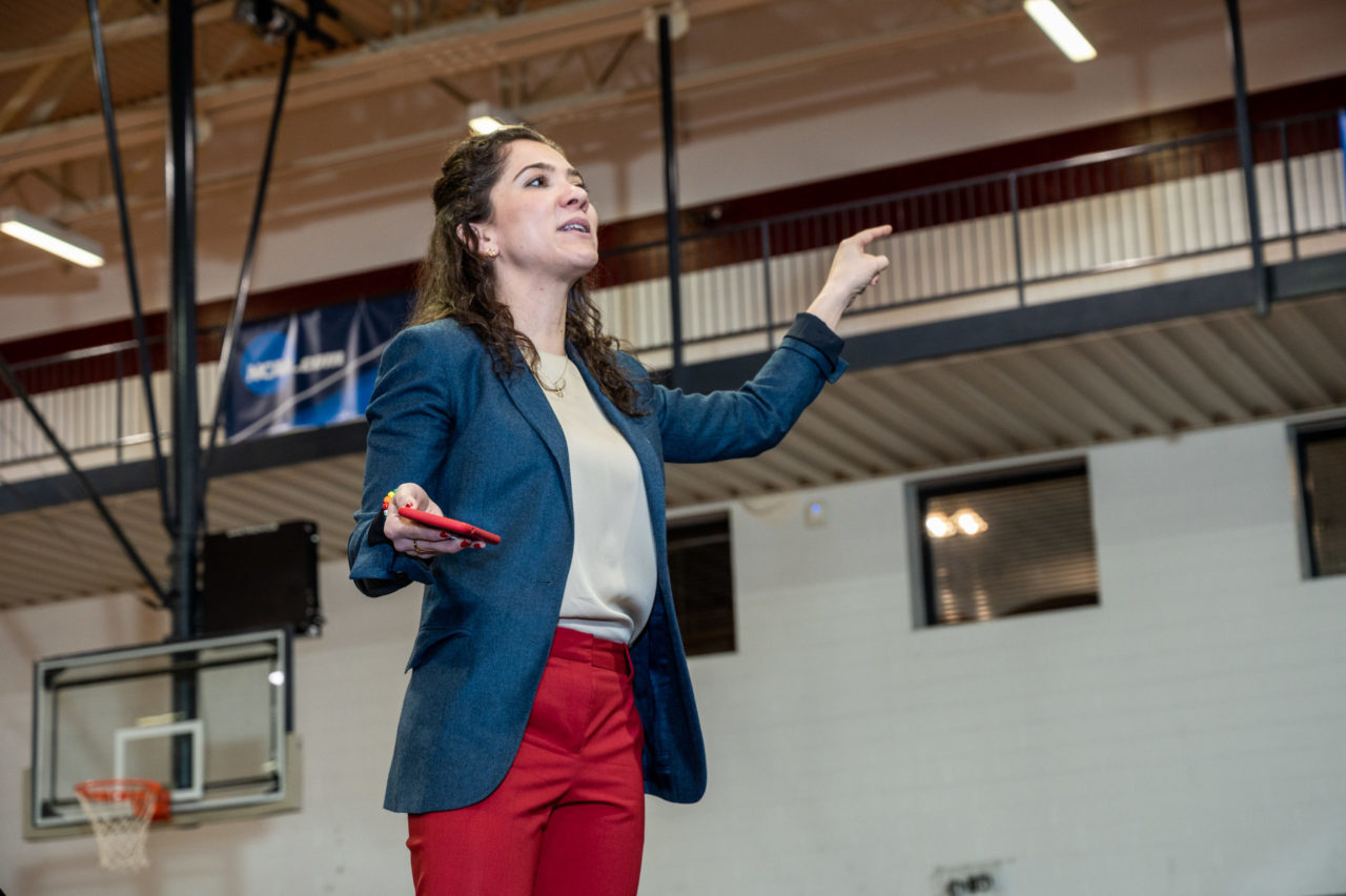 Female stands with arms outstretched, speaking to an audience outside of the frame.