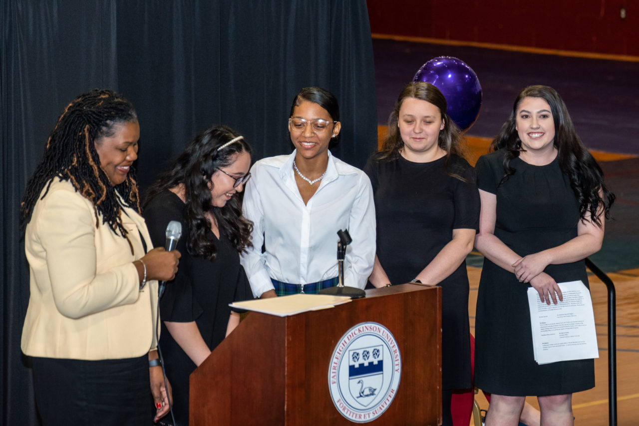 Five females stand side by side behind a podium, looking down and smiling.