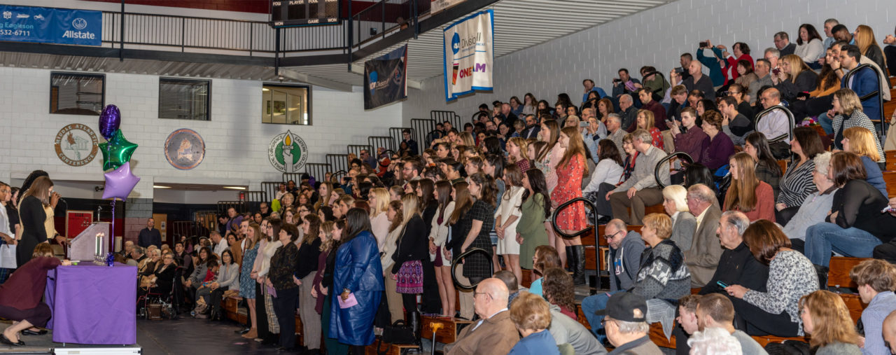 A crowd of students and their families stands in the bleachers facing the stage.