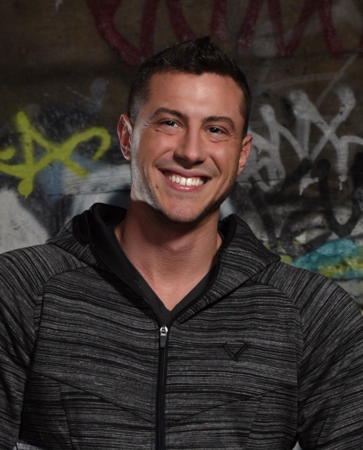 A male in a dark grey hooded sweatshirt smiles at the camera.