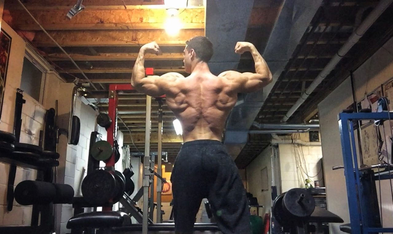 Male bodybuilder stands and faces away from the camera, with arms flexing at ninety degree angles from his torso.