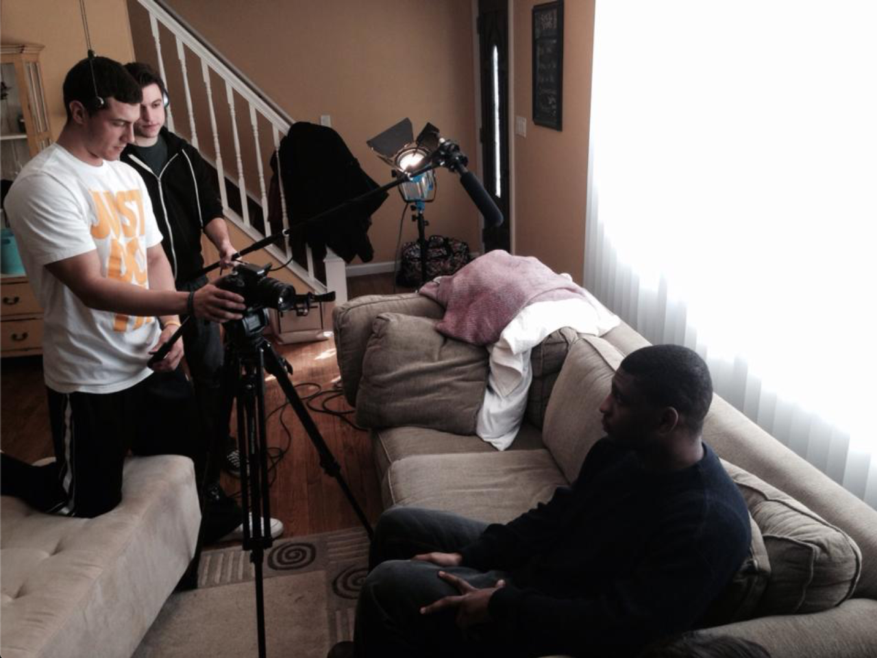 Two males stand behind a camera and a boom microphone as a third male sits on a sofa to their right.