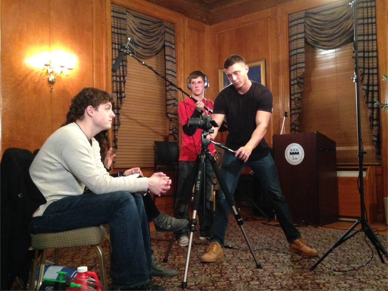 A male sits on a chair facing right while two other males stand and position a boom microphone and a camera for filming.