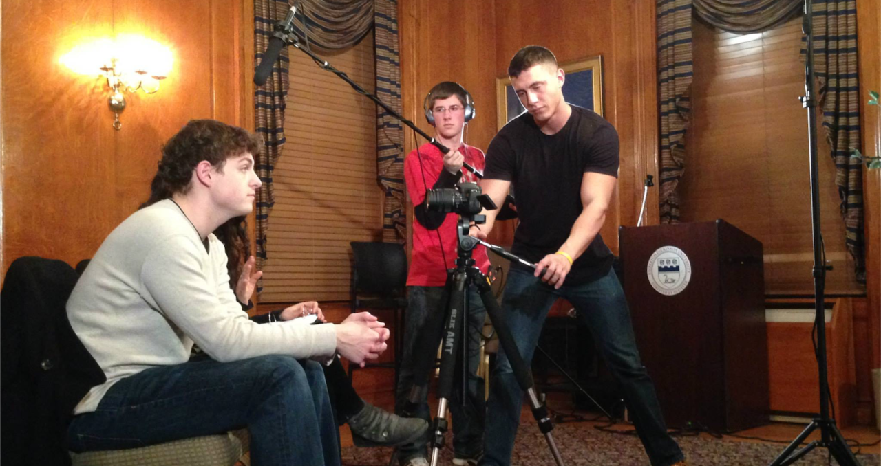 A male sits on a chair facing right while two other males stand and position a boom microphone and a camera for filming.