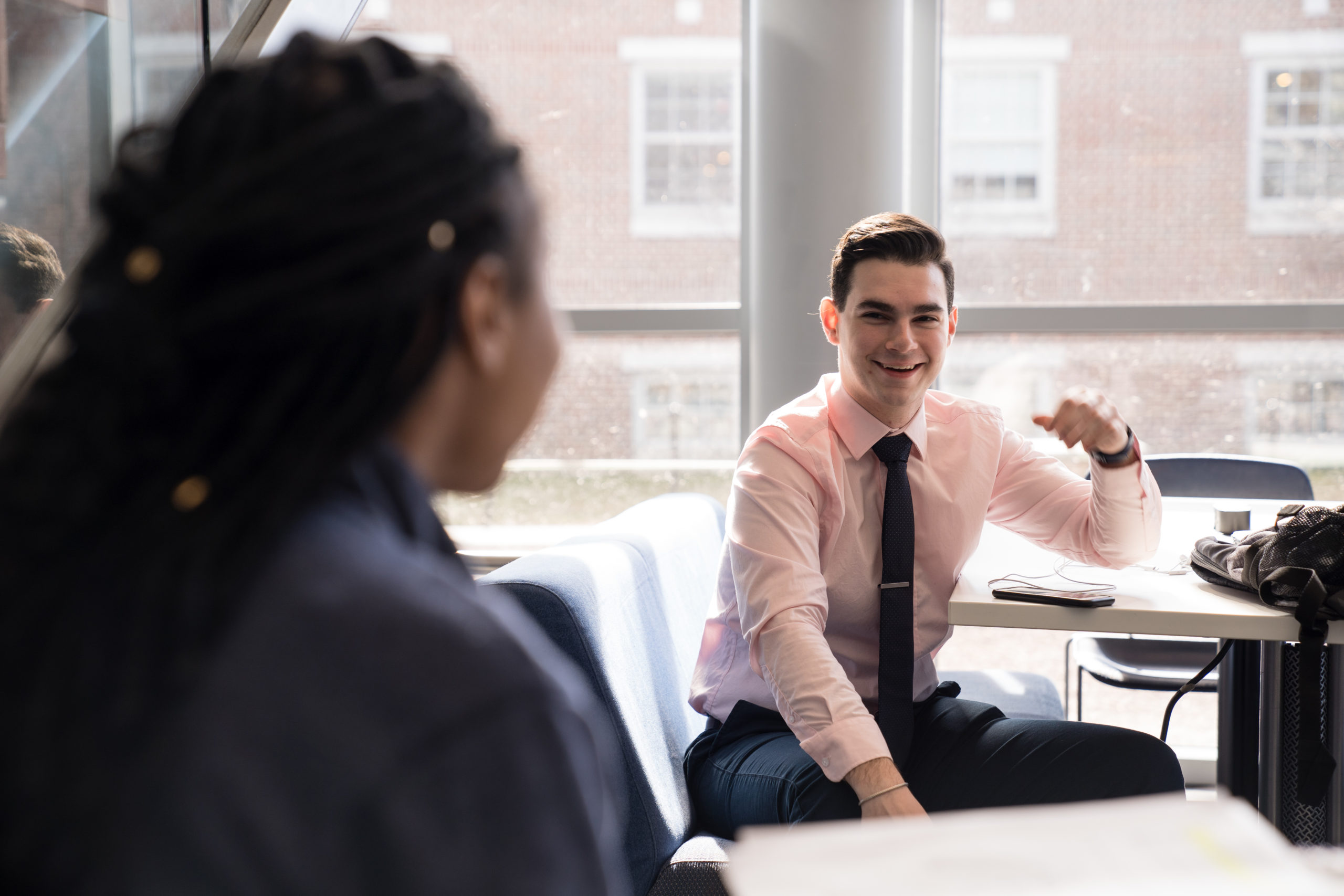 A student in a shirt and tie sits and smiles. He is across from someone who's back of head is visible on the left.