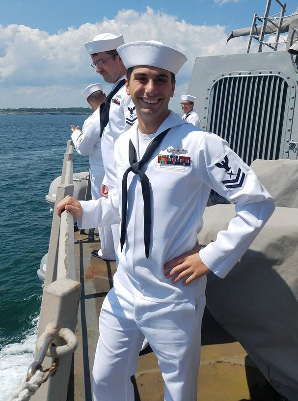 Dillon Pontes, a naval officer, stands on the deck of a ship in his uniform, with his right hand resting on the railing overlooking the ocean. Another sailor stands behind him.
