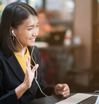 A woman with headphones on waves at someone she is video chatting with on a laptop