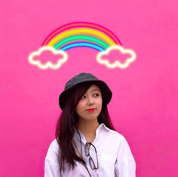 A young woman stands in front of a bright pink wall, with a rainbow above.
