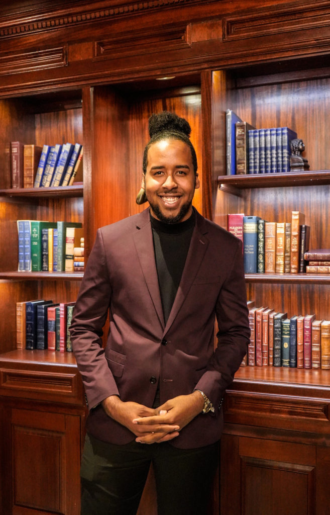A young man stands in front of a bookcase.