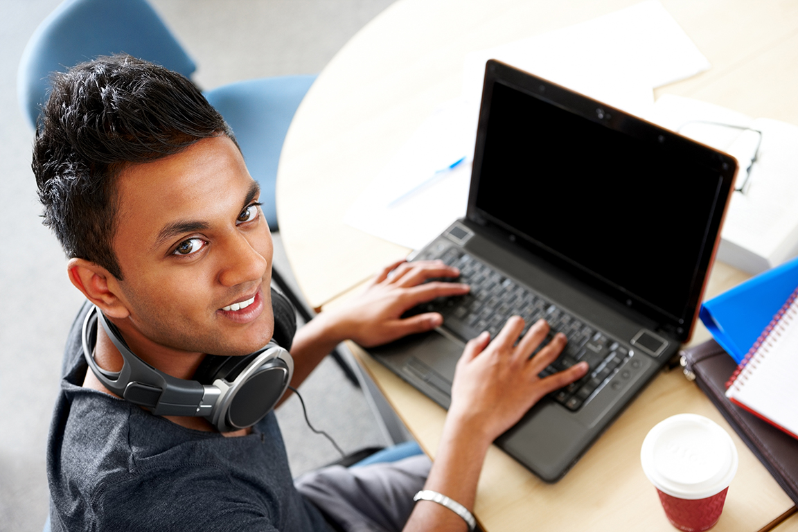 Student using laptop at a desk. Headphones are around his neck.