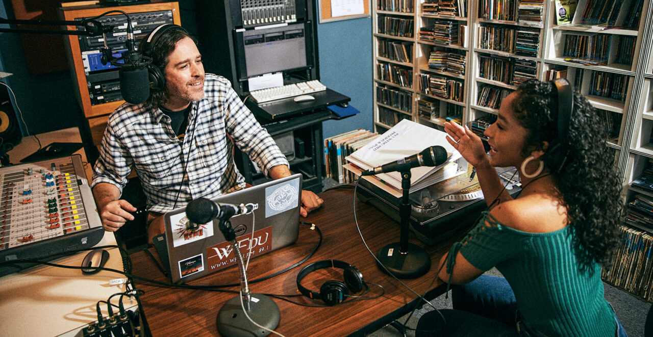 A man and a woman sit behind microphones in a radio station recording studio.