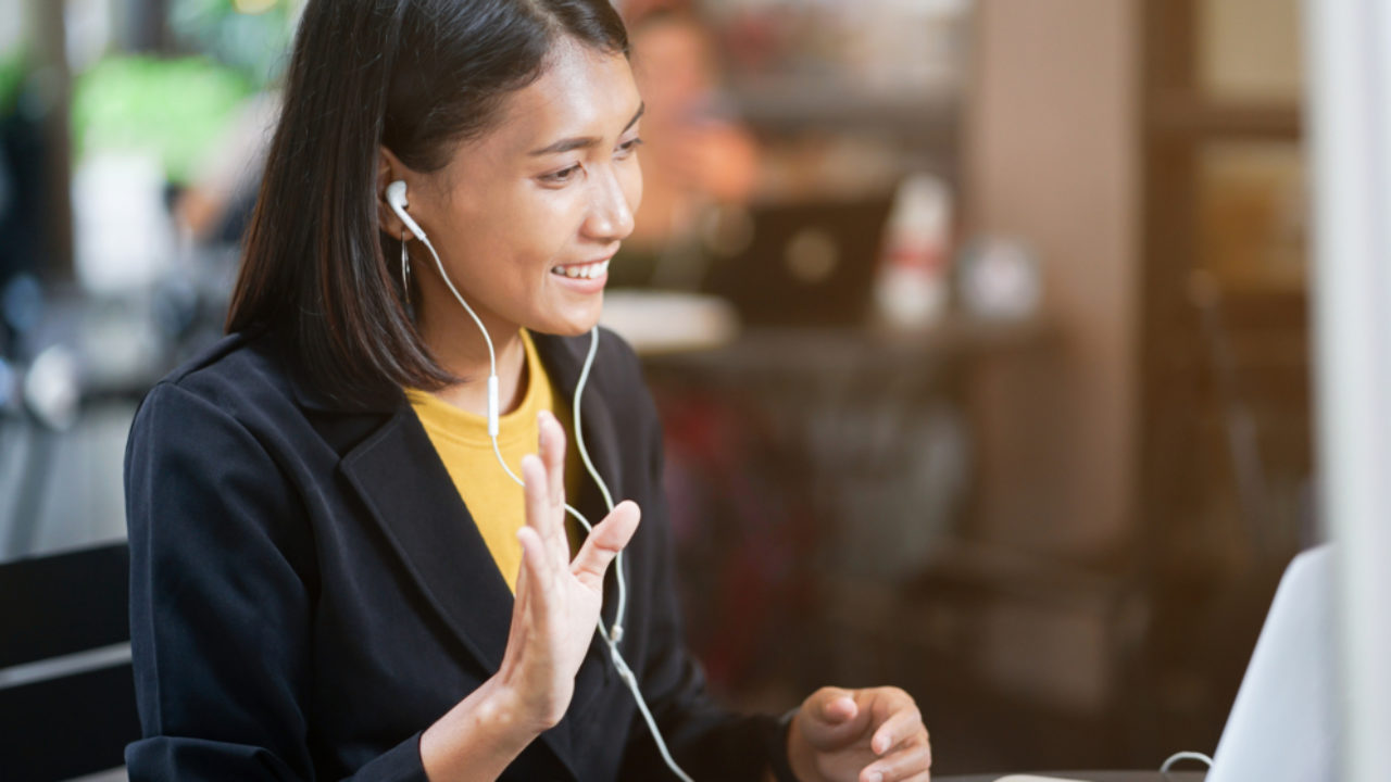 A woman with headphones on waves at someone she is video chatting with on a laptop