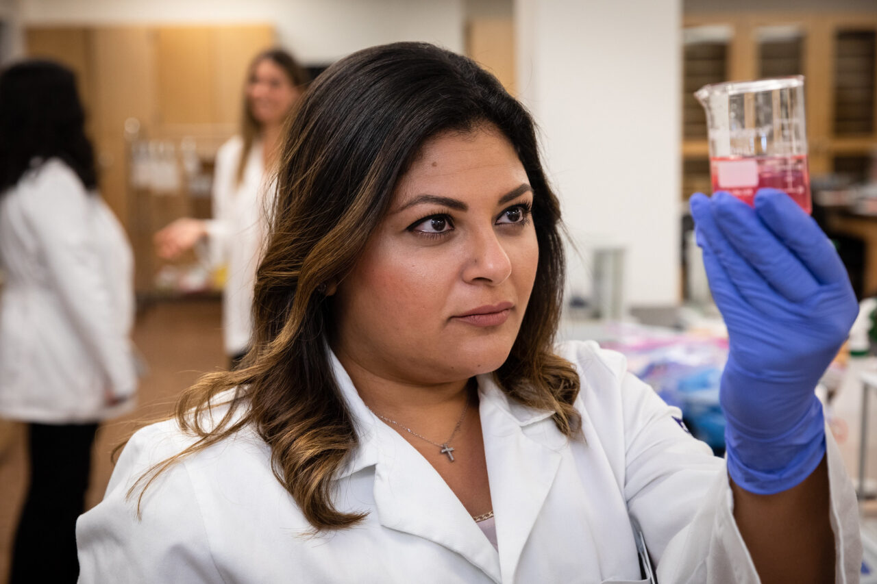 a student wears a white coat and holds a beaker. she is in a lab.