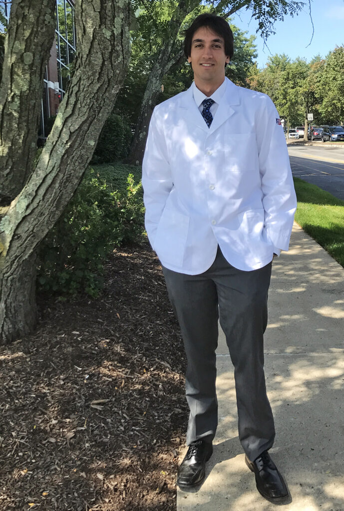 A young man wearing a white coat stands outside of a building.