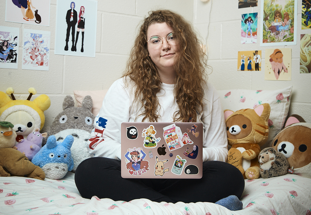 A young woman sits cross-legged on her dorm bed, holding her laptop.