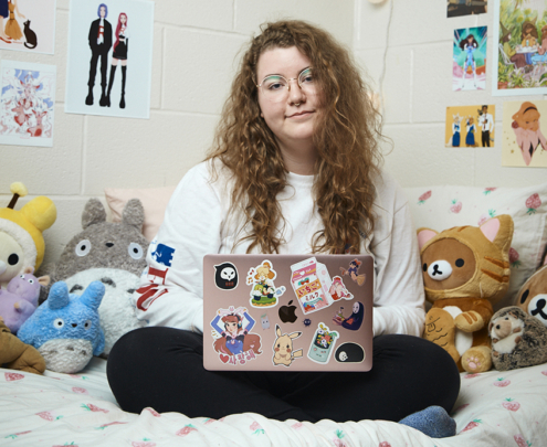 A young woman sits cross-legged on her dorm bed, holding her laptop.