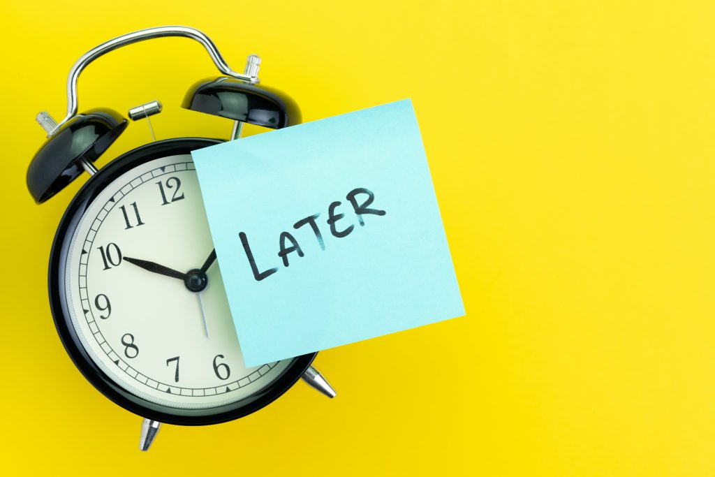 Sticky post with handwriting the word Later stick on alarm clock on solid yellow background with copy space using as procrastination, self discipline or laziness concept.