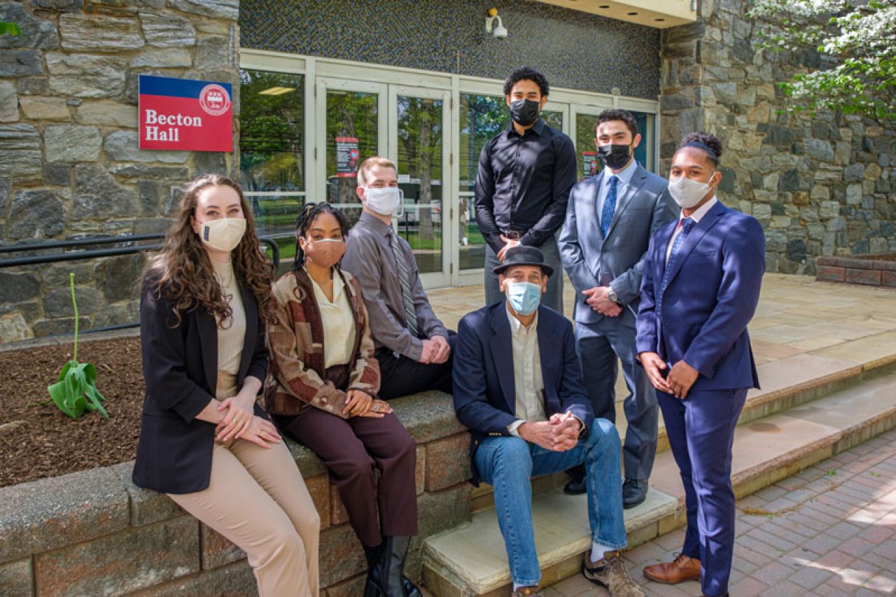 A group of students wearing masks sit and stand on the steps outside of Becton Hall.