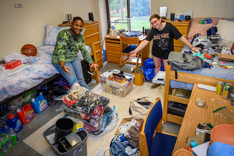 Two young women stand, arms out wide, in the middle of a messy dorm as they unpack.