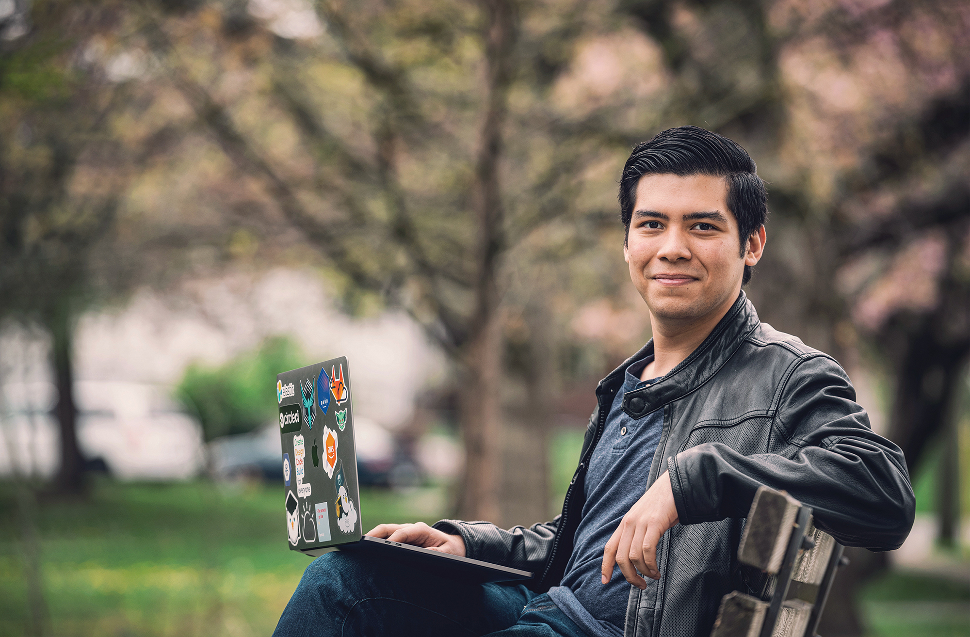 A young man sits on a park bench, laptop open on his knee.