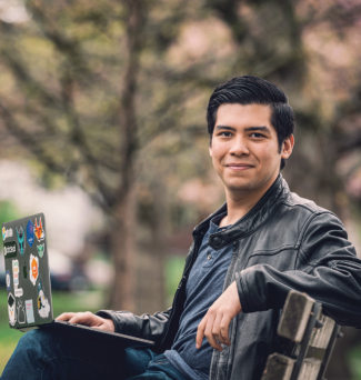 A young man sits on a park bench, laptop open on his knee.