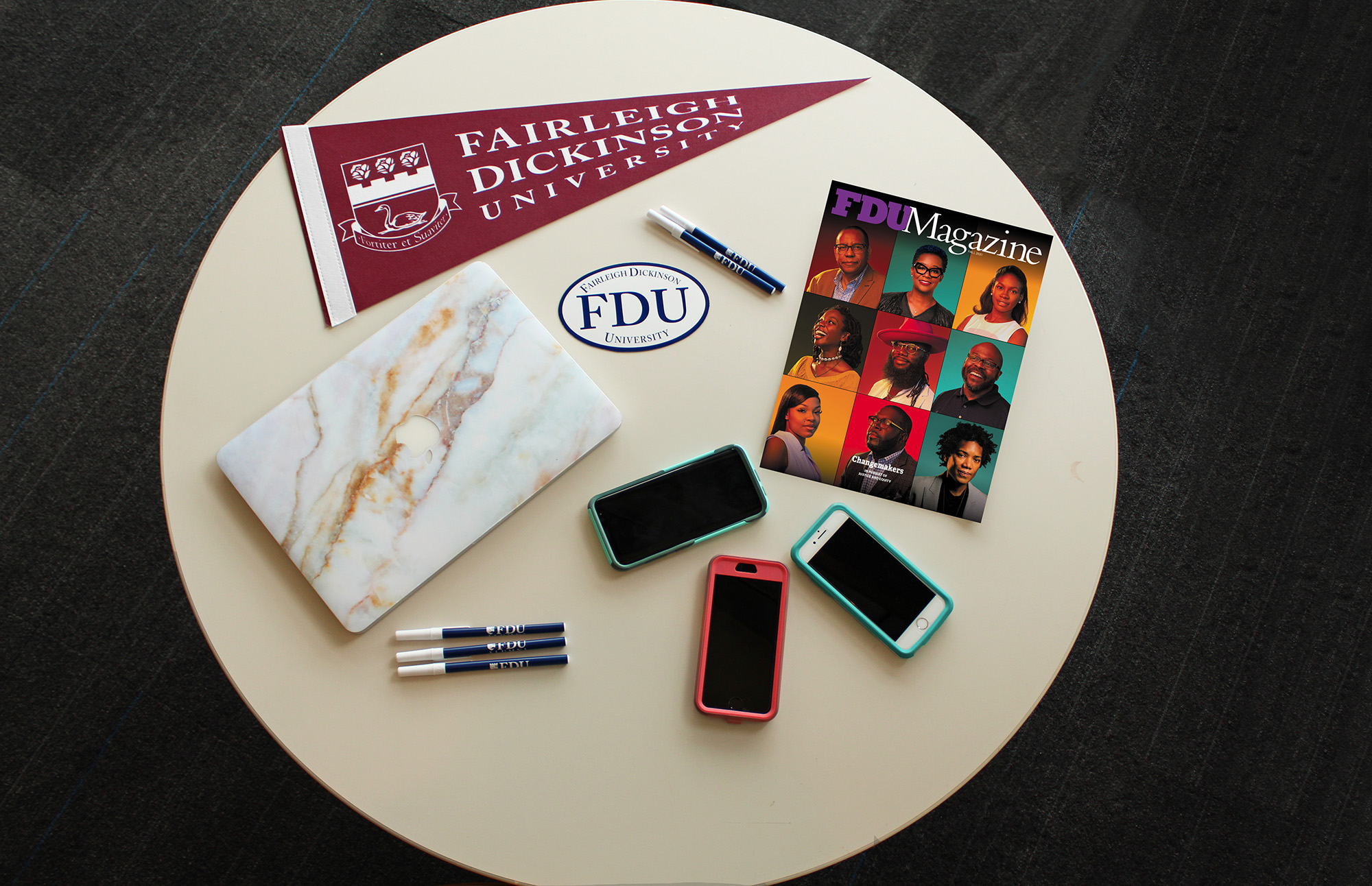 Still life of a table with an FDU pennant, a copy of FDU Magazine, three smartphones, a laptop and pens.