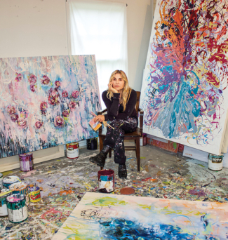 A female artist sits in her studio surrounded by her paintings and artwork.