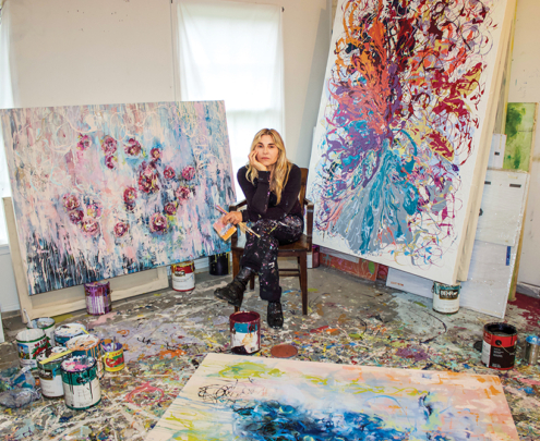 A female artist sits in her studio surrounded by her paintings and artwork.