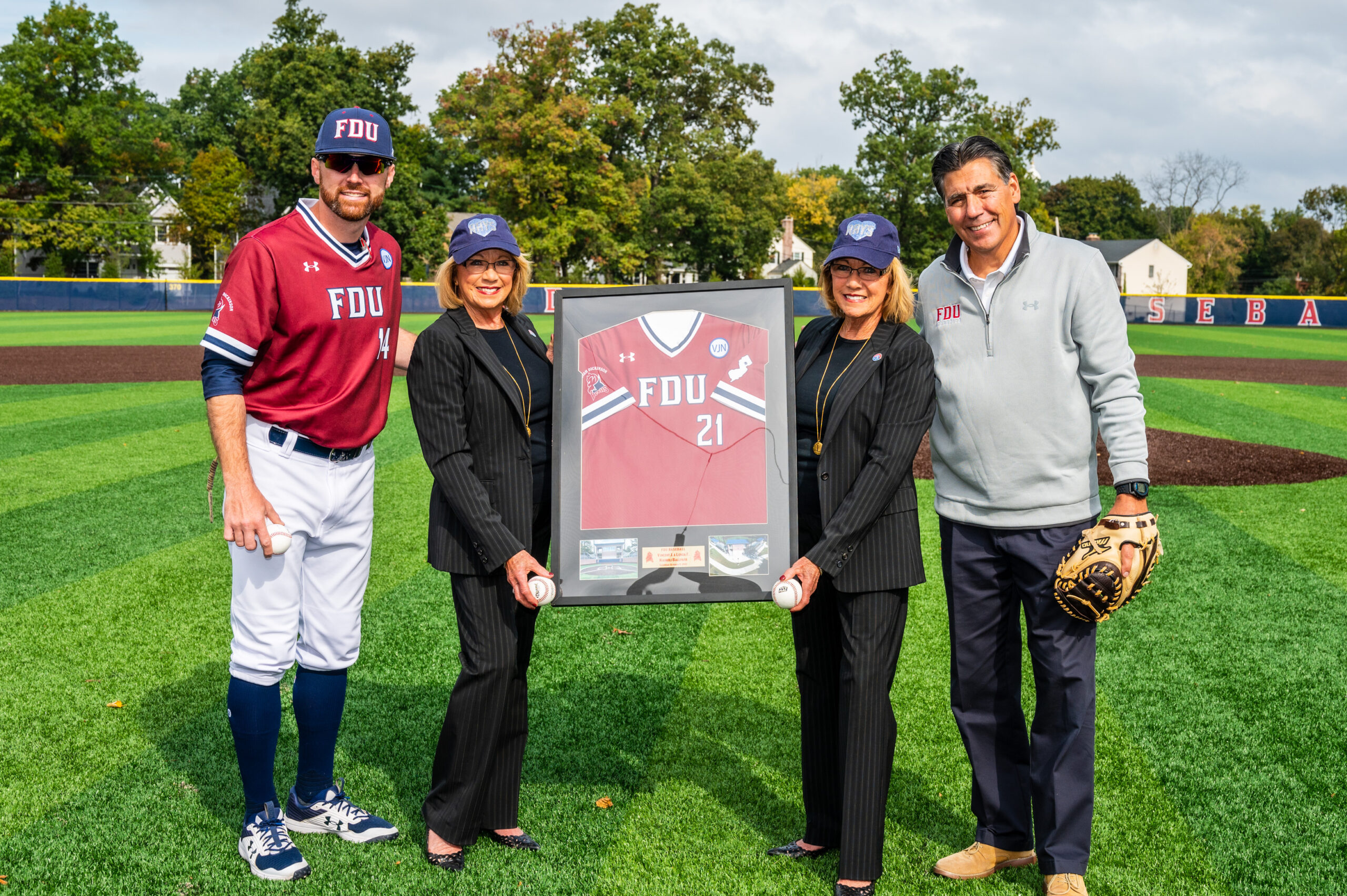 Two men and two women pose for a photo as the women hold a framed FDU baseball jersey.