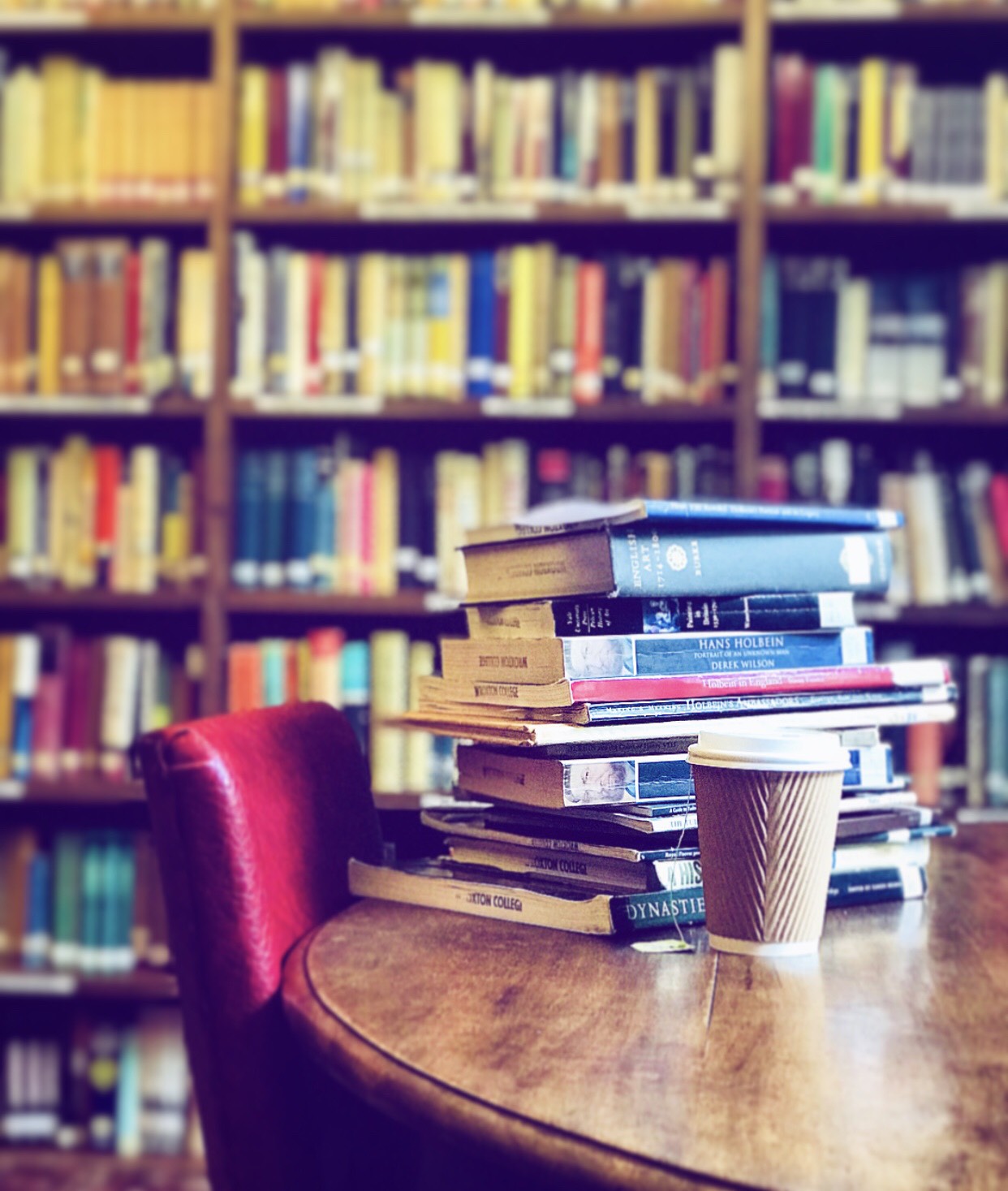 Textbooks stacked on a table, next to a disposable coffee cup and a row of bookshelves behind.