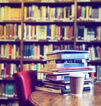 Textbooks stacked on a table, next to a disposable coffee cup and a row of bookshelves behind.