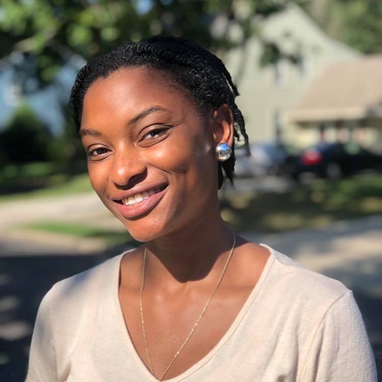 Portrait of a young woman, standing outside in the sunlight. She's smiling with a tilt of the head.