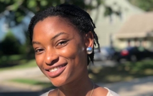 Portrait of a young woman, standing outside in the sunlight. She's smiling with a tilt of the head.