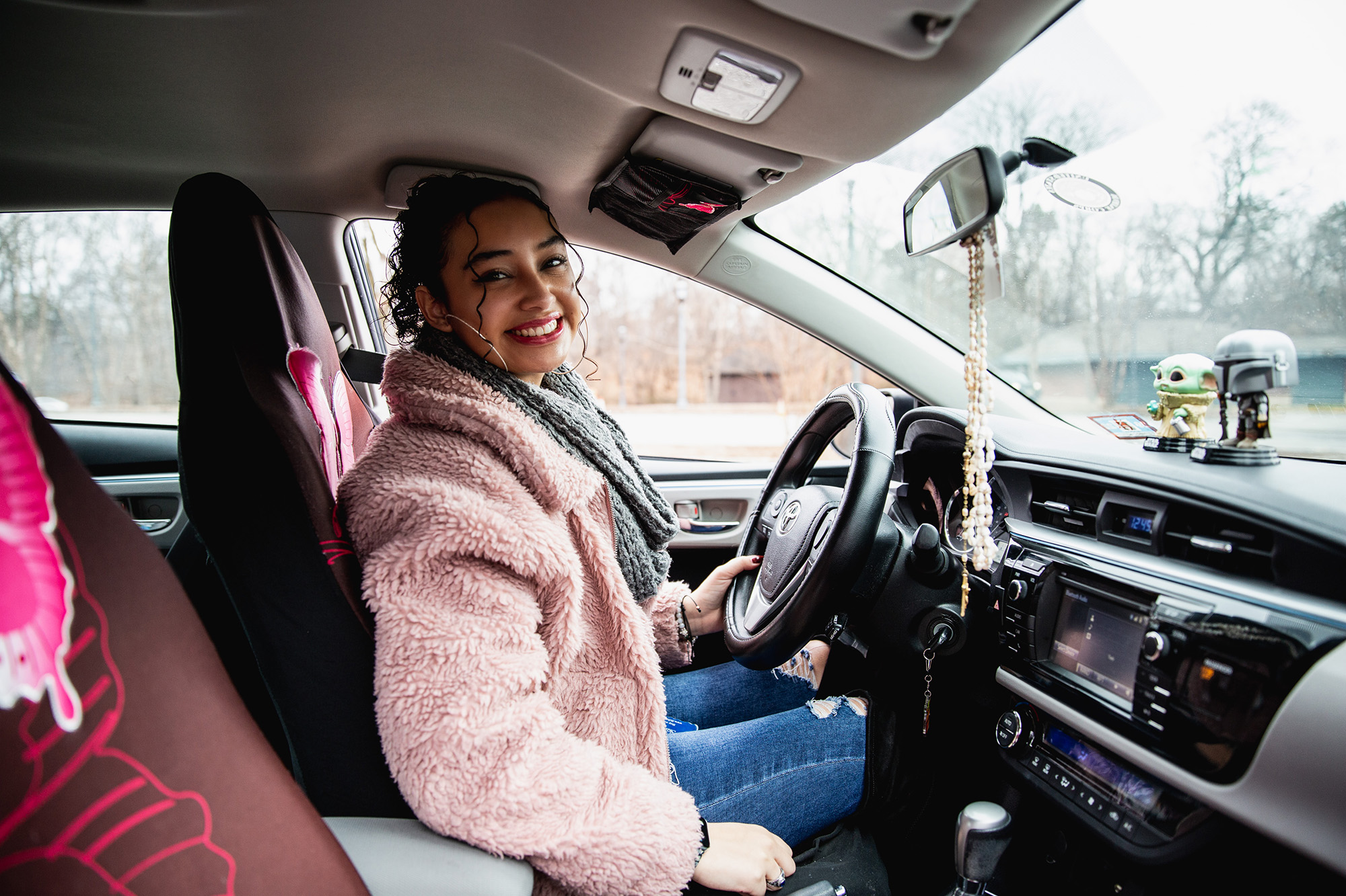 A young woman wearing a fuzzy pink coat sits in the driver's seat of a car, smiling.