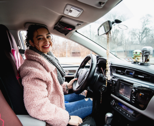A young woman wearing a fuzzy pink coat sits in the driver's seat of a car, smiling.