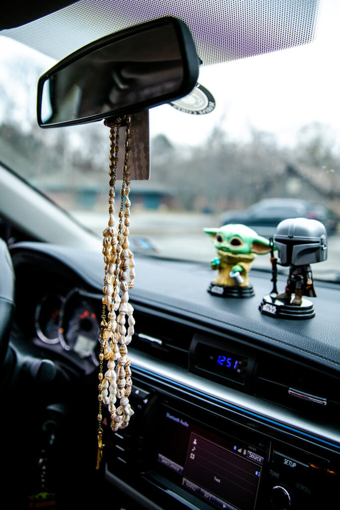 Two Star Wars figurines sit on a car dashboard. A rosary and seashells hang from the rearview mirror. 