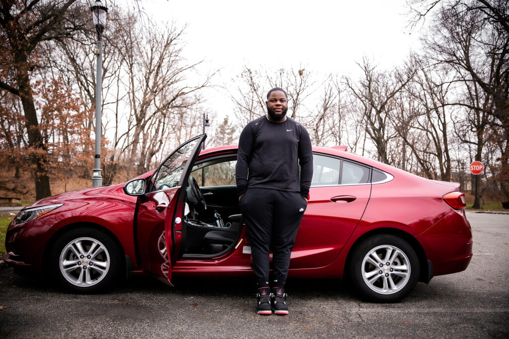 A young man stands against his red car.