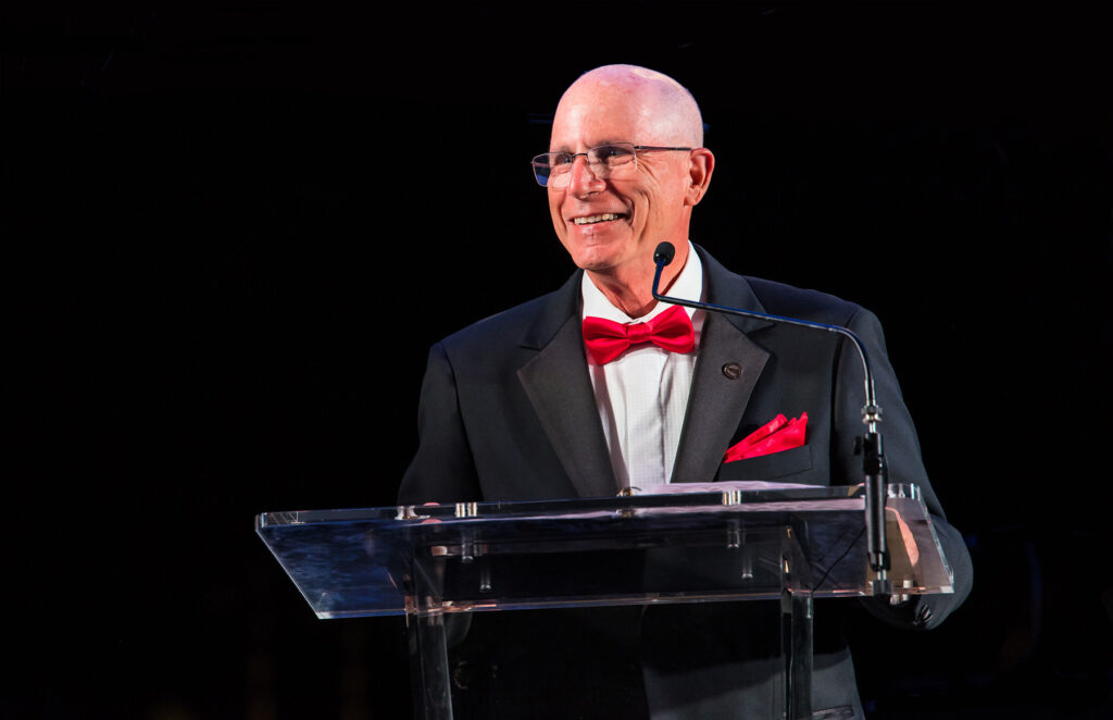 A man wearing a tuxedo with a red bowtie speaks at a podium.