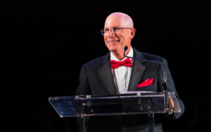 A man wearing a tuxedo with a red bowtie speaks at a podium.