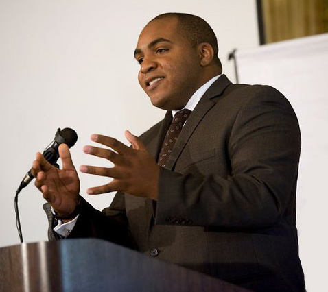 A man in a suit stands, speaking at a podium.