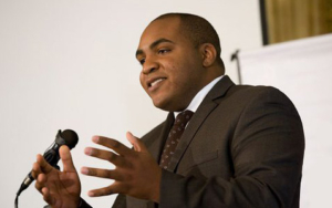 A man in a suit stands, speaking at a podium.