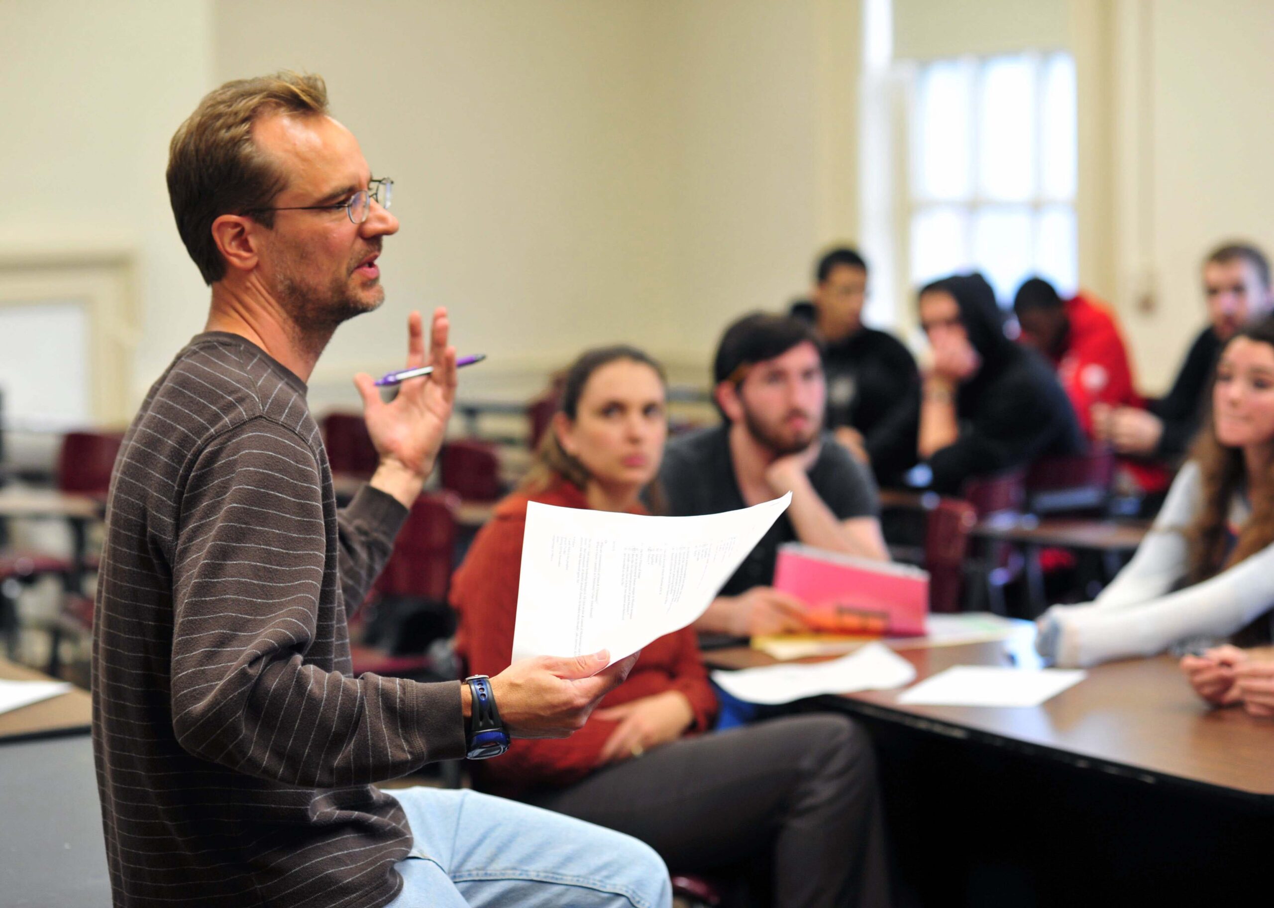 A male professor sits on the edge of a desk, holding a piece of paper, with a class of students before him.