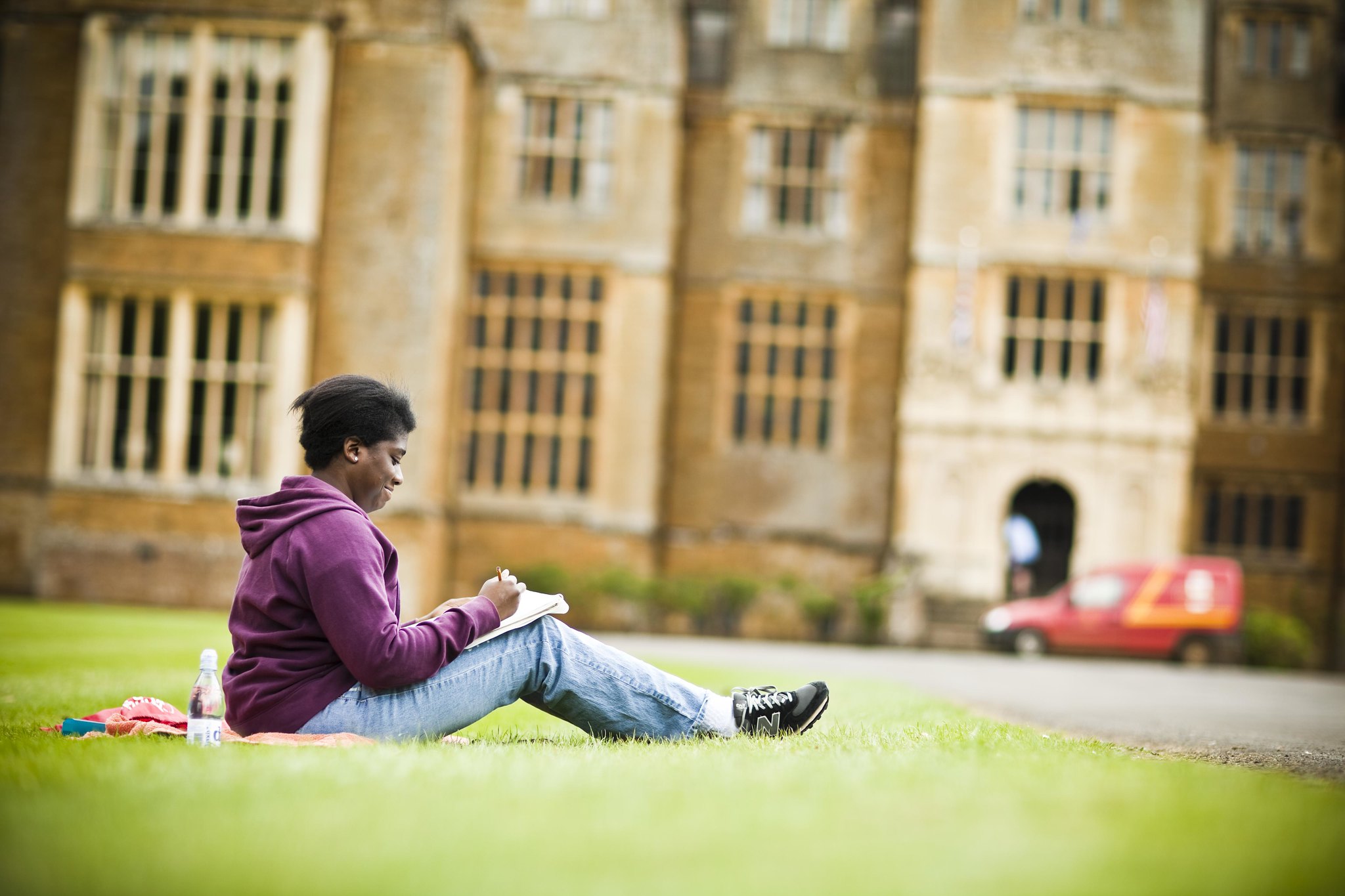A student sits on the grass in front of a historic brick building, engrossed in reading or studying while enjoying a sunny day on campus.