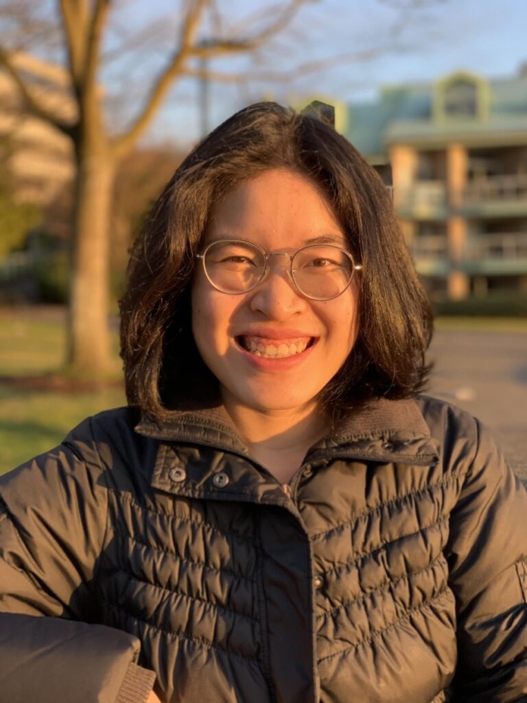Portrait of Anh Pham, wearing glasses and a coat on a sunny day.