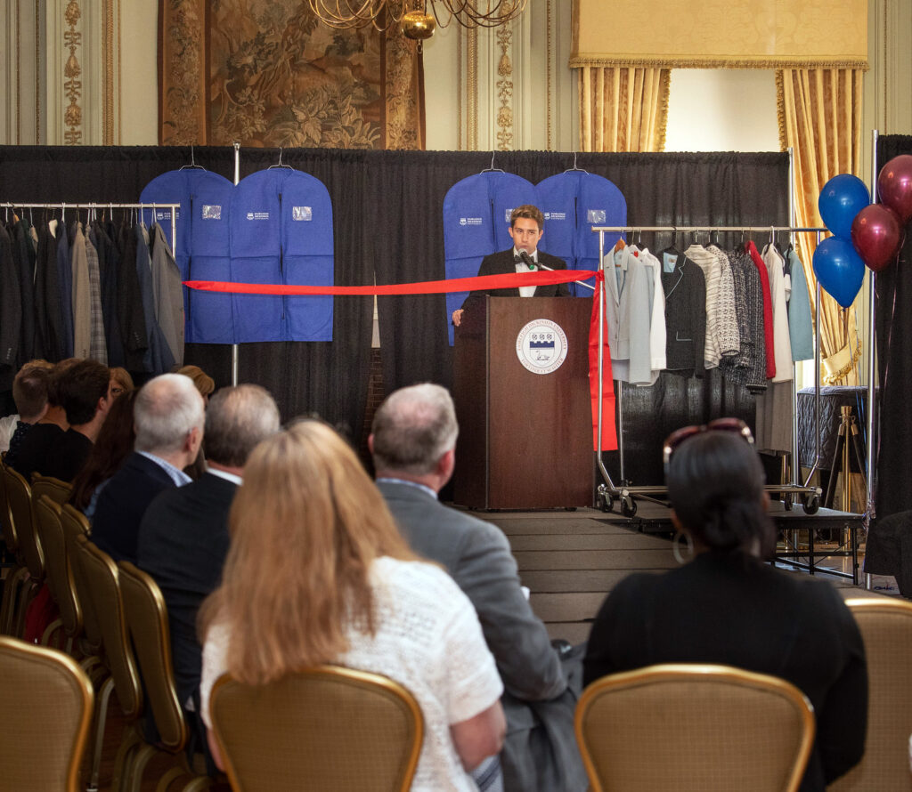 A young man in a tuxedo stands a podium, on a fashion show runway, delivering remarks.