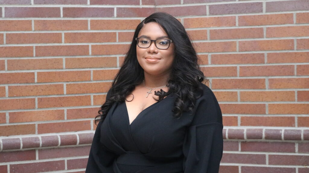 Lisa-Monet Jefferson, wearing glasses and a black shirt, stands in front of a brick wall.