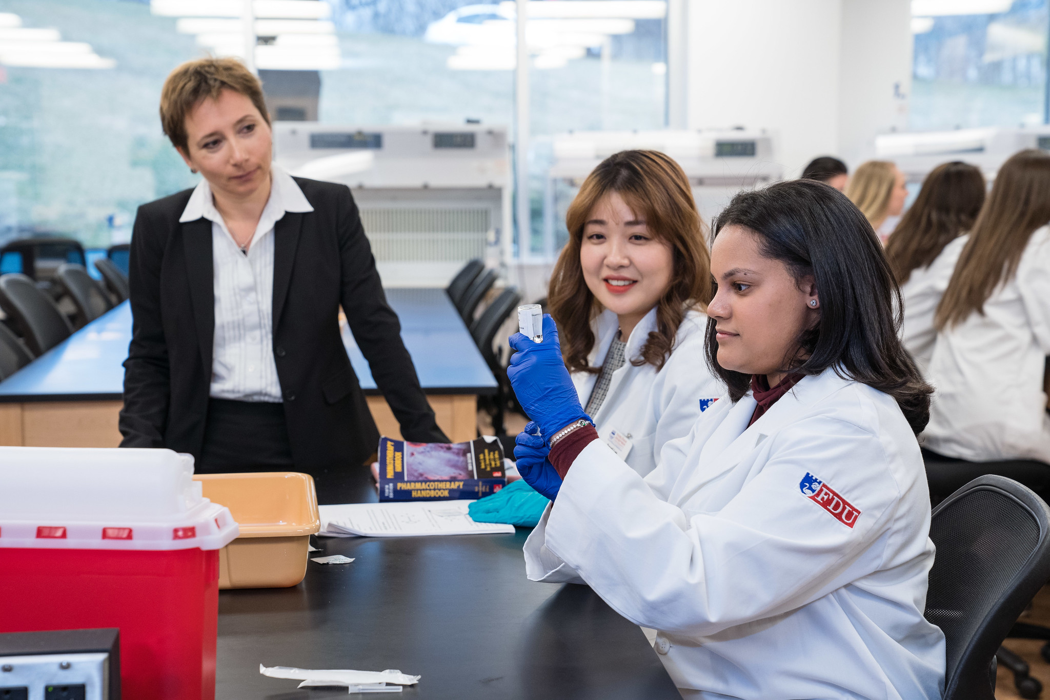 A female professor watches a pharmacy student wearing a lab coat practice drawing medication into a syringe.